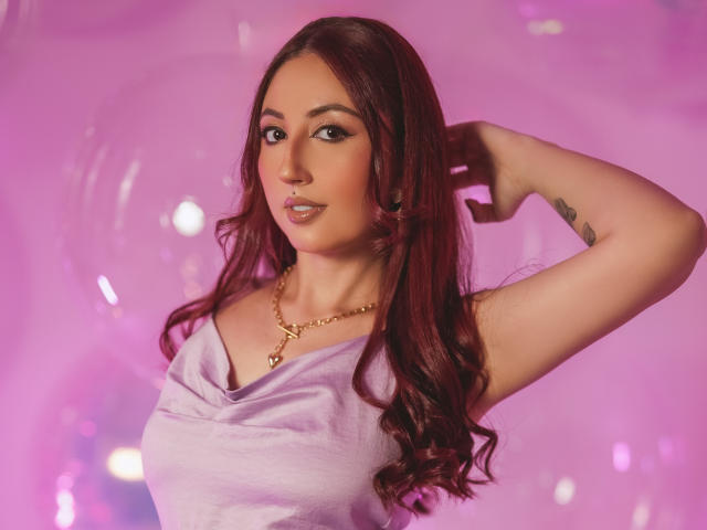 EmiilyWatson with long red hair, wearing a purple top, gold necklace, and looking at the camera with a slight smile, hand in hair, showing a tattoo on her arm.