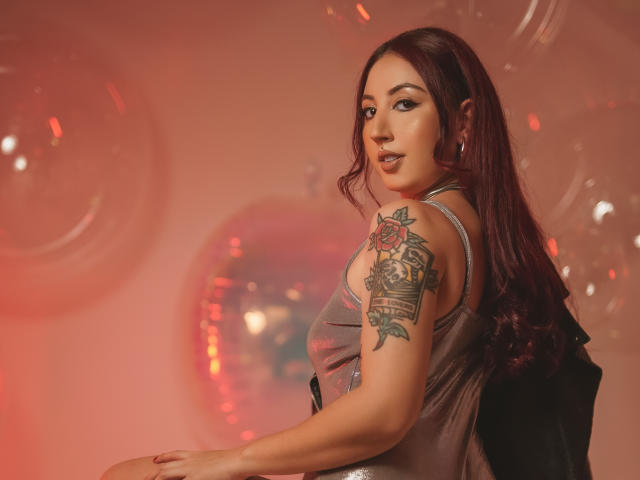 EmiilyWatson with long red hair, looking over her shoulder, wearing a silver dress, showing a colorful tattoo on her right arm, red lights in the background.