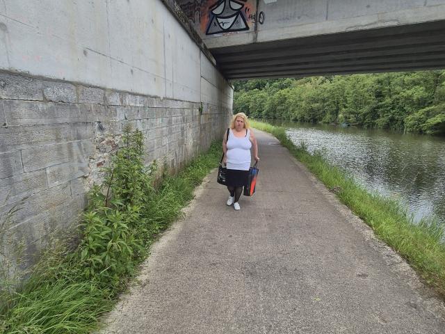 StephanieLeo69, chubby blonde with white tank top, black pants, and white sneakers, walks on concrete path under bridge, carrying red and black bag, with green grass and river.