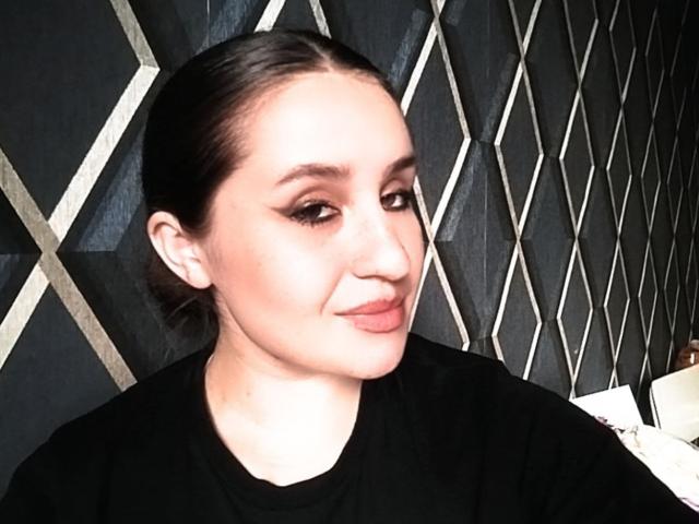 Drayana with slicked-back brown hair, black eyeliner, and pink lips, wearing a black shirt, smiling softly. Her ass is not visible.
