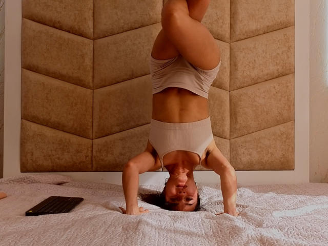 LucyySky is doing a headstand on a bed, wearing beige shorts and a tank top, with her tits and ass slightly visible, in a cozy, brown-upholstered room.