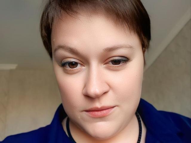 YourGoddessX has a short brown pixie cut, fair skin, brown eyes with black eyeliner, and is wearing a blue shirt, with a neutral expression on her face.