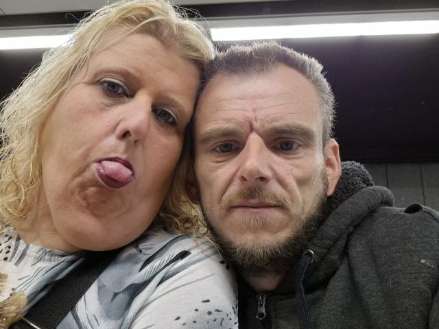 StephanieLeo69 with blonde, wavy hair and pouty pink tongue, leaning on a bearded man with gray hoodie, both in a subway car. His mustache is slightly curled.