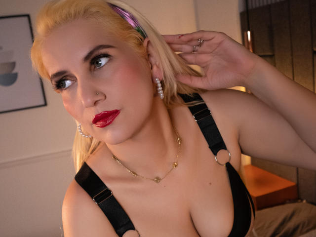 GiuliaAddams has blonde hair, blue eyes, red lipstick, and is wearing a black bra with straps, showing her large tits. She's touching her hair with a ringed hand.