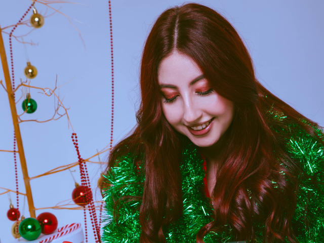 EmiilyWatson with long red hair, smiling, wearing green tinsel, looking down, red and green Christmas ornaments on bare tree, white background, soft light, cute face.