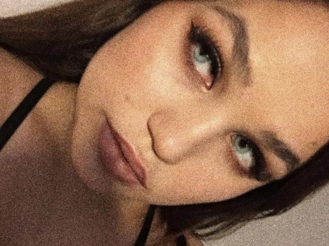 AnnyMarryah has a pouty face with full lips, green eyes, and brown eyebrows. She's wearing a black strappy top, and her brown hair is partially visible.