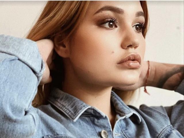 AnnyMarryah's face with light brown skin, full lips, and a small beauty mark on her cheek. She's wearing a denim shirt, hands behind her head, brown eyes looking up.