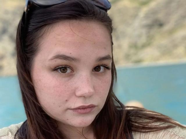 AnnyMarryah has brown eyes, straight dark brown hair, and a light skin tone with small freckles. She's wearing sunglasses on her head and a beige top.