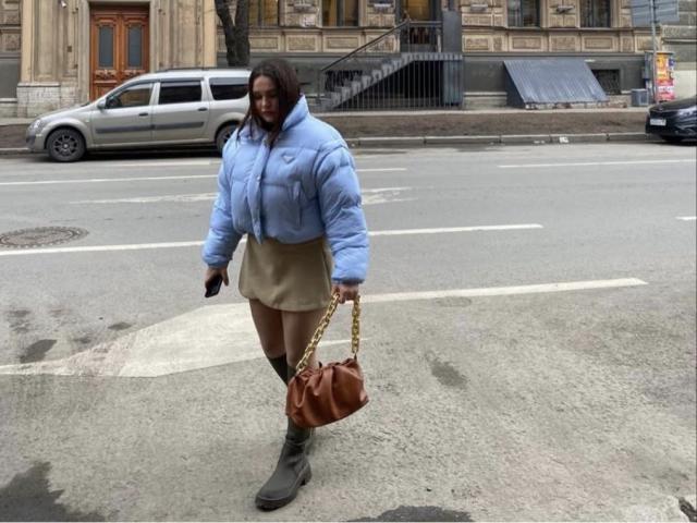 AnnyMarryah with blue puffy jacket, beige skirt, grey boots, holding brown bag with gold chain, standing on street, holding black phone, in front of silver SUV.