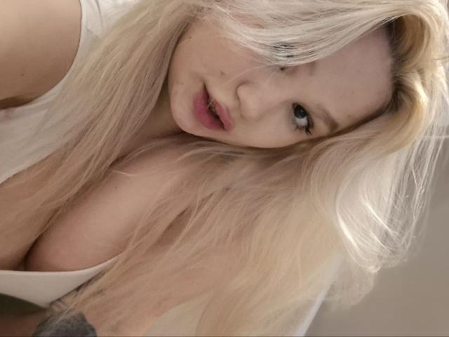 AnnyMarryah has long blonde hair, pale skin, and pink lips, looking at the camera with a slight pout. Her white top shows large, cleavage-filled tits.