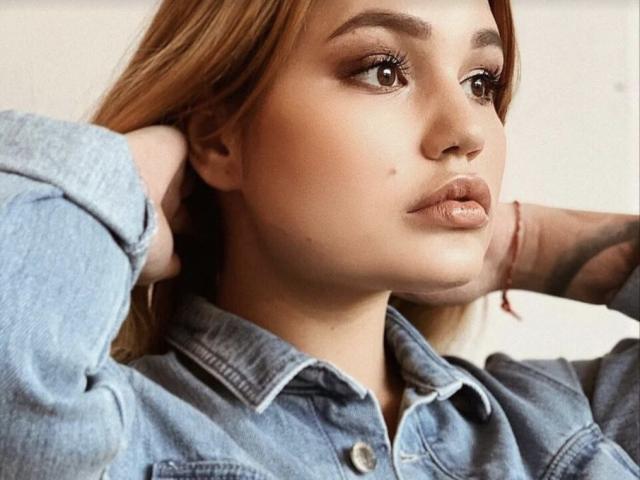 AnnyMarryah with pouty lips, brown eyes, and light brown hair, wearing a denim shirt, hands in her hair, subtle tattoo on her right arm. Soft, natural light.