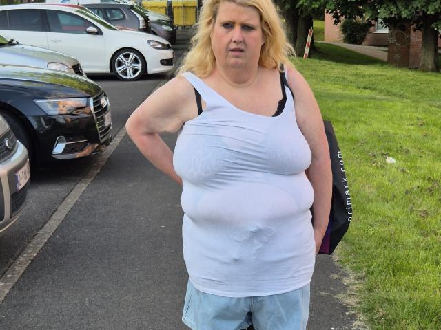 A chubby StephanieLeo69 with blonde hair stands in a parking lot, wearing a wet white tank top that shows her large tits and blue jeans, with hands on hips.