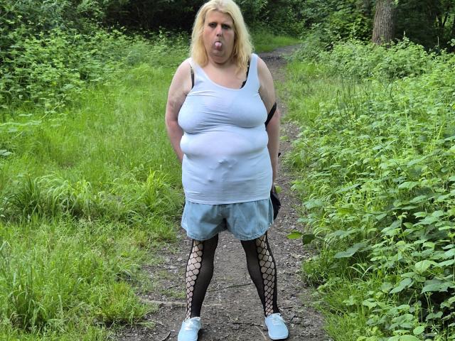 StephanieLeo69 stands on a dirt path, tongue out, wearing a tight white shirt over her big tits, blue shorts, fishnet stockings, and white shoes. She's chubby and blonde.