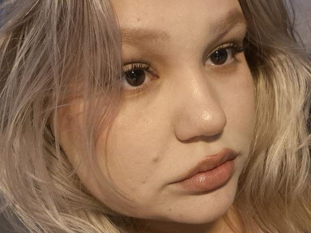 AnnyMarryah's face with light skin, brown eyes, and slightly parted pink lips. She has messy blonde and gray hair, a small beauty mark on her right cheek.