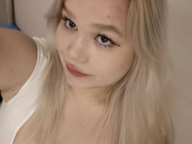 AnnyMarryah has blonde hair, blue eyes, and a small beauty mark on her left cheek. She's wearing a white shirt with a slightly open neckline.