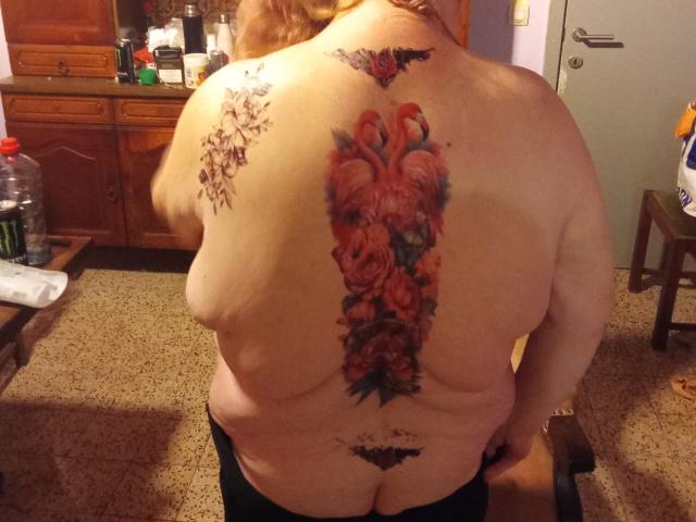 StephanieLeo69's back tattoo featuring a large, colorful, red and orange bird with roses, centered on her pale, chubby, topless body, showing a lower black tattoo and some skin.