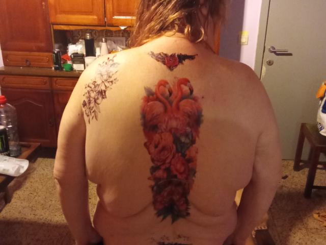 StephanieLeo69 has a large, colorful tattoo of roses on her back, with additional smaller tattoos on her right shoulder, and is standing naked in a kitchen.