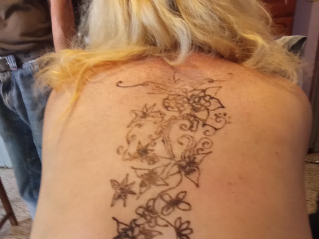 StephanieLeo69's back tattoo featuring a black floral design with swirls and leaves, positioned vertically, showing her blonde hair in the top half of the image.