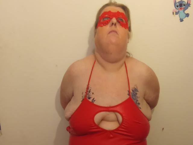 StephanieLeo69, chubby with large tits, wearing a red leather halter top and matching red lace mask, puffs out her chest, tattoos on her shoulders, standing against a plain wall.