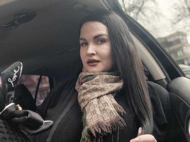 GeorgiaLerox with long dark hair, wearing a black coat and brown patterned scarf, holding a black glove with white embroidery, looking at the camera with a serious expression.