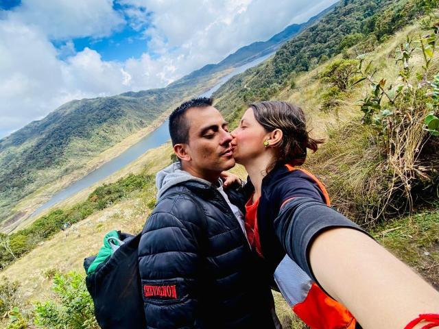 LucianSins, a man with short black hair and a medium build, wearing a black puffy jacket, kisses LucianSins, a woman with brown hair in a ponytail, wearing a black and red jacket. LucianSins takes a selfie with his right arm. LucianSins has a green backpack. The background shows a mountainous landscape with a river and green foliage. Bright, cloudy sky. LucianSins' face is close to LucianSins' cheek. LucianSins' right ear has a gold hoop earring. LucianSins' red jacket contrasts with LucianSins' black jacket. LucianSins' face is slightly turned to the right. LucianSins' left arm is partially visible.