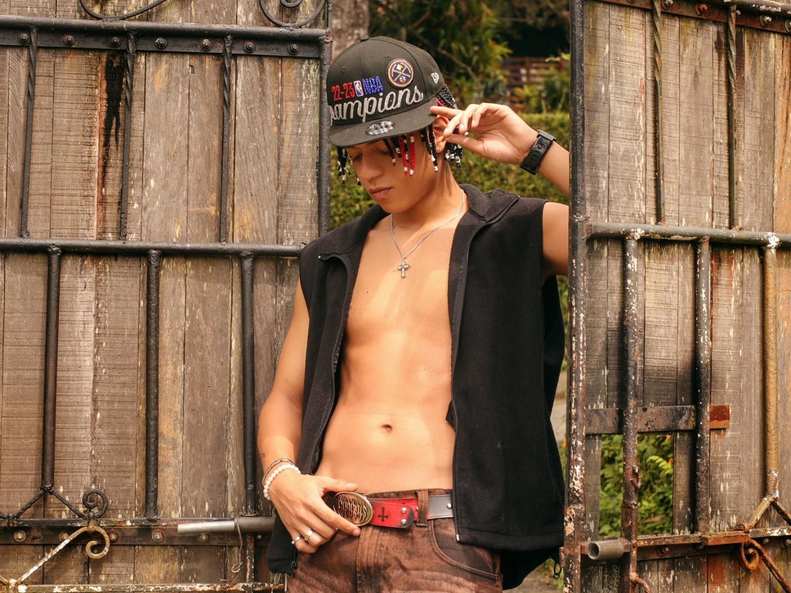 MarshallSpencer with a bare, toned, tan torso, wearing a black sleeveless vest, brown pants, red belt, and a black "Pompin's" hat, standing in a rusty wooden gate.