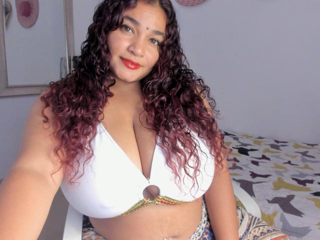 DarsshaHara, con cabello castaño rizado, lápiz labial rojo, un top de bikini blanco que muestra un busto grande, y un anillo de oro, sonriendo a la cámara, sentada en una cama con una funda estampada..