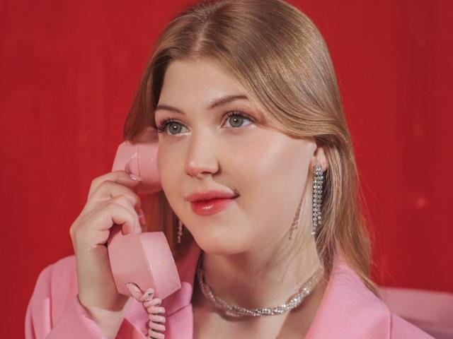 StellaLane, with light brown hair and green eyes, holds a pink vintage phone to her ear. She has fair skin, red lipstick, and wears a silver chain necklace and dangling earrings. Her pink jacket contrasts with the red background. She looks slightly upward, with a relaxed expression. The image focuses on her upper body and face. The pink phone and jacket create a cohesive color theme. The photo is clear and sharply focused.