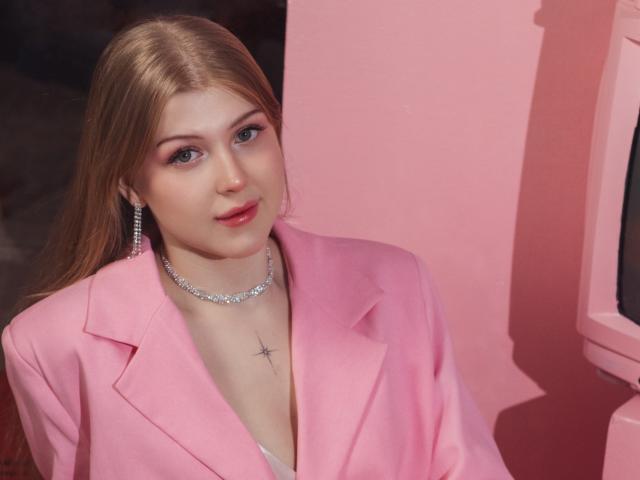 StellaLane with long blonde hair, pink blazer, silver necklace, and earrings, showing a cross tattoo on her chest, looking cute with red lipstick.