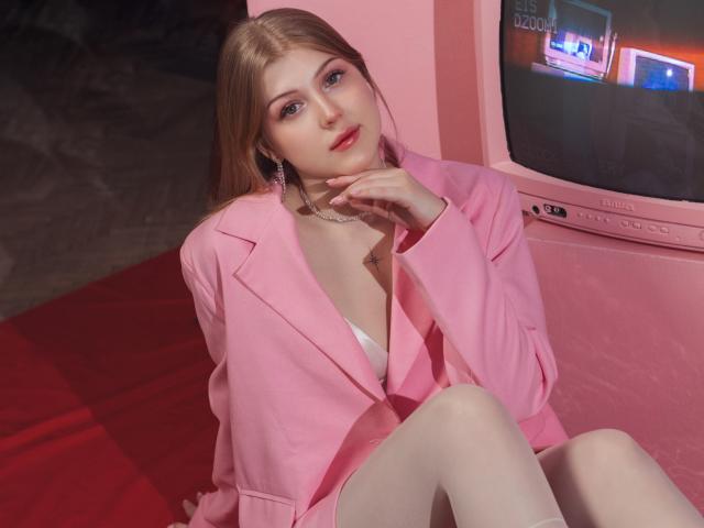 StellaLane with blonde hair, pink blazer, white bra, and panties, sitting on red carpet, legs crossed, hand on neck, looking at camera, pink wall, TV screen in background.