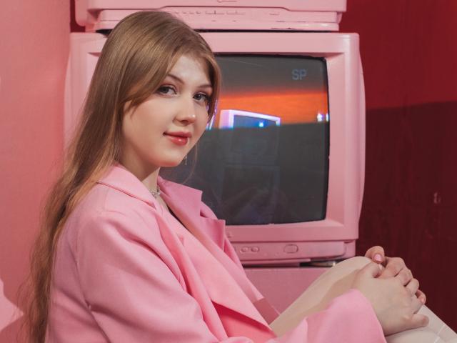 StellaLane with long brown hair, pink blazer, and pink TV in the background, sitting with hands on lap, smiling slightly, red-pink room, light skin, pink lips.