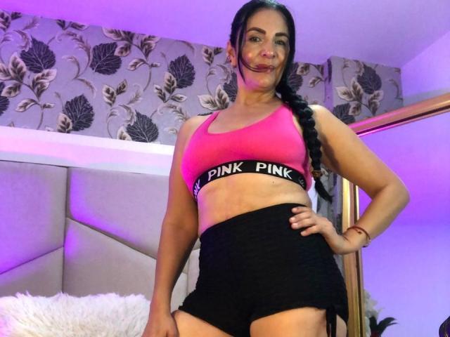 AlizonMoreti with long black braid, pink sports bra, black shorts, pink underwear band, standing, hand on hip, smiling, purple and white lighting, bedroom setting.