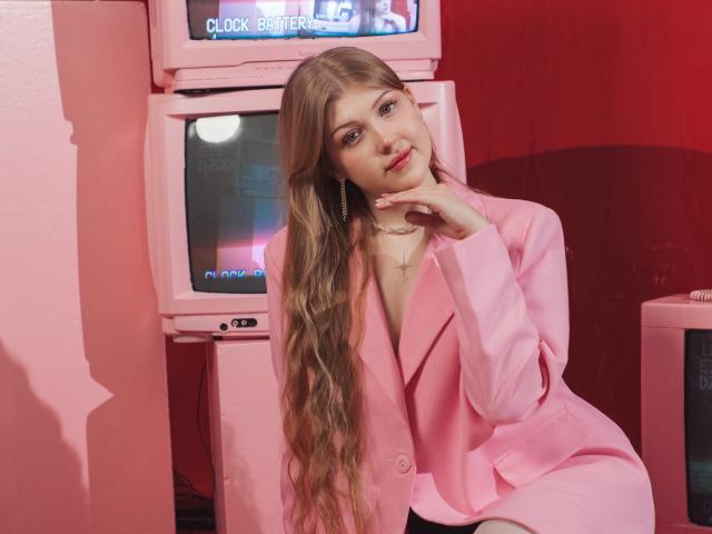 StellaLane with long, wavy blonde hair, pink blazer, and necklace, sitting in front of pink TVs, hand on chin, smiling, pink wall, red shadow.