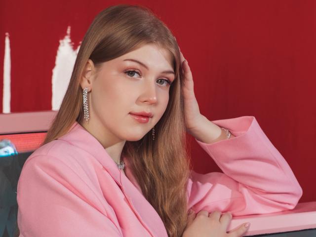 StellaLane has long, straight brown hair and wears a pink blazer with a silver necklace. She's leaning on a pink surface with one hand in her hair. Her makeup is subtle with pink eyeshadow and glossy pink lips. She has light skin and wears dangling silver earrings. The red background contrasts with her pink outfit. The image is a photograph with a soft, even lighting that emphasizes her relaxed, confident expression.