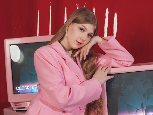 StellaLane with long brown hair, pink blazer, and white necklace, leaning on a pink TV, looking at the camera with a slight smile, right hand on her cheek.