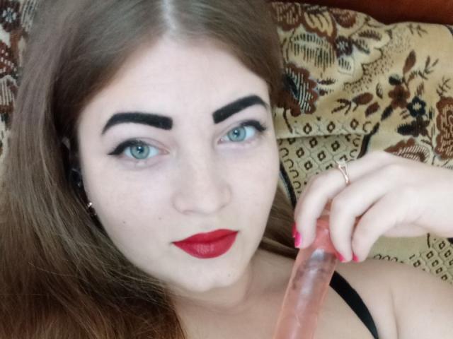 ElizabethHottie with fair skin and blue eyes, has dark eyebrows and red lipstick. She holds a translucent pink dildo with her right hand, which has red-painted nails. Her straight brown hair falls to the sides. She wears a black strap over her shoulder. The close-up focuses on her face and the dildo. The background is a patterned fabric. Her expression is neutral. She has a ring on her right index finger. The image is sharply focused on her face and hand.