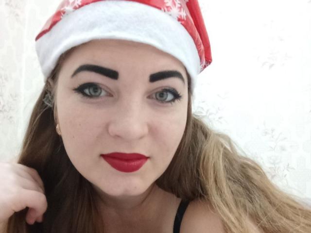 ElizabethHottie with red Santa hat, thick black eyebrows, green eyes, red lipstick, long brown hair, and gold earrings, holding her hair. Boobs visible, black strap showing.