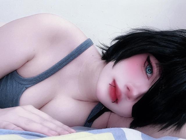 AngelaRumiee with black hair, pale skin, blue eyes, pink blush, gray tank top, biting lower lip, lying on bed, hand on blanket, visible tits, slightly parted lips.