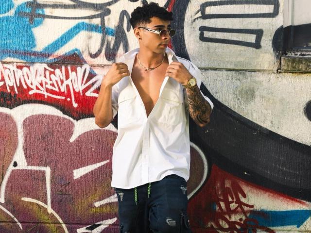 AlexZT with short black hair, tattoos on right arm, wearing a white unbuttoned shirt, glasses, and dark pants, standing against graffiti-covered wall, holding shirt edges.