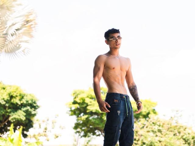 Shirtless AlexZT with a tattooed arm, wearing dark ripped jeans and sunglasses, standing outside, sunlight highlighting his toned, muscular, tan body and short dark hair.