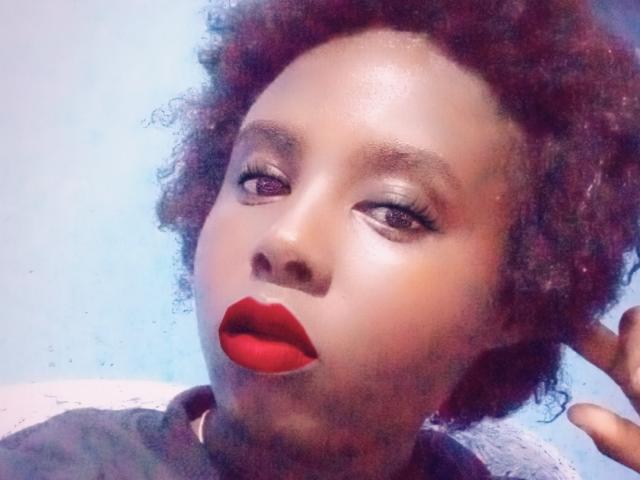 Close-up of Mamonah with dark brown skin, curly red hair, and glossy eyes. She has bold red lipstick and subtle eye makeup. Her expression is slightly open-mouthed, with one hand near her head. The lighting is soft, highlighting her smooth skin and the texture of her hair. The image has a slight blue tint. Mamonah's face is centered, occupying most of the frame. The composition emphasizes her facial features and red lipstick. The overall style is modern and slightly surreal.