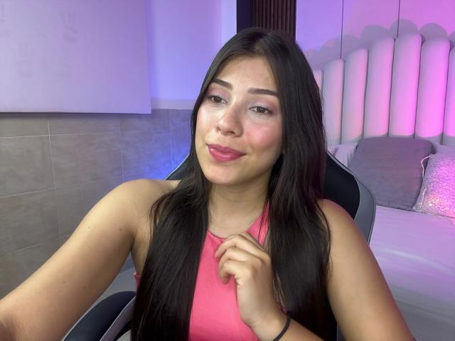 IsabelaPretty with long black hair, pink tank top, slight smile, and pink lipstick, sitting in a chair, holding her shirt, with a cute, sexy, and attractive appearance.