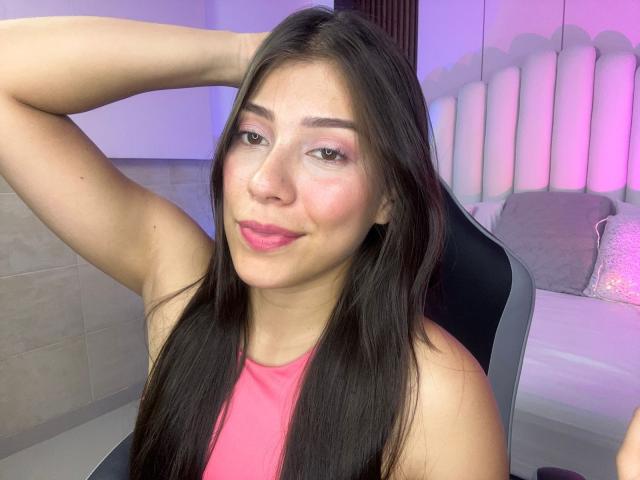 IsabelaPretty with long black hair, pink lipstick, and pink eyeshadow, smiling, arm raised, wearing a pink tank top, sitting in a black chair.