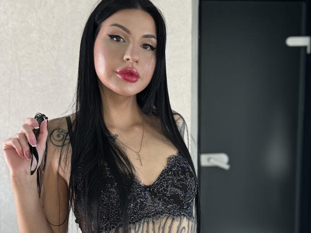 AuraXo with long black hair, wearing a black lace top, holding black straps, has tattoos on her arm and chest, red lips, and a cross necklace, looking at camera.