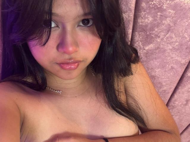 Mei69 met glanzende, roze lippen, donker, golvend haar en een zilveren ketting, die haar kleine borsten met één hand bedekt, voor een achtergrond van roze fluweel..