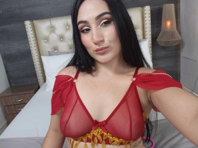 HiilaryDolce with long black hair, wearing a red sheer bralette showing her small tits, and a yellow patterned top, smiling in a bedroom with a tufted headboard.