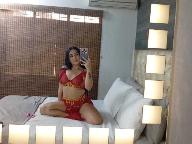 HiilaryDolce sits on a bed in red lingerie with yellow chains, holding a phone, her pussy barely visible, with a mirror reflecting her, in a brightly lit room.