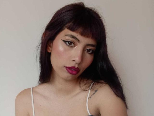 AndreaVargass with dark purple hair, pink lipstick, black eyeliner, nose ring, white spaghetti strap top, small tattoo on right shoulder, looking at camera, pale skin, subtle blush.