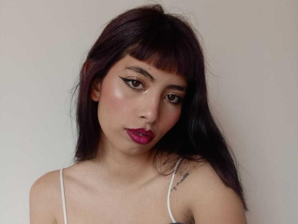 AndreaVargass with dark purple hair, pink lipstick, black eyeliner, nose ring, white spaghetti strap top, small tattoo on right shoulder, looking at camera, pale skin, subtle blush.