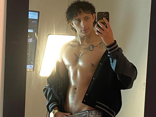 JayTaylor, avec des cheveux bouclés, torse nu, veste noire, tatouages sur la poitrine, tenant un téléphone, autoportrait devant un miroir, montrant des abdominaux musclés, main dans le pantalon, regardant la caméra, pièce avec une source de lumière..